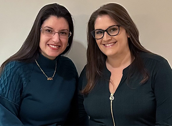 Two women with long dark hair wearing glasses and dark colored sweaters smiling at the camera.