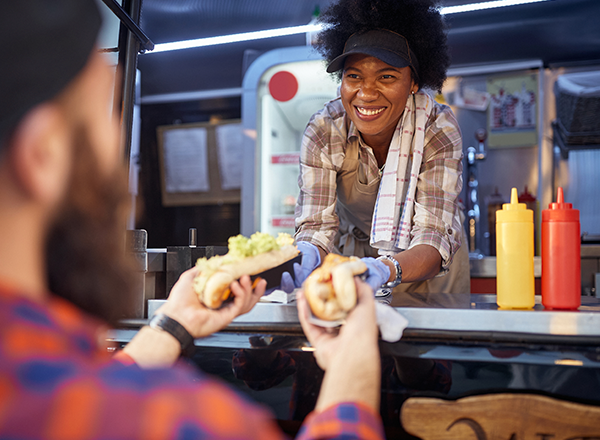 Smiling woman inside food truck passing two sandwiches to a customer.