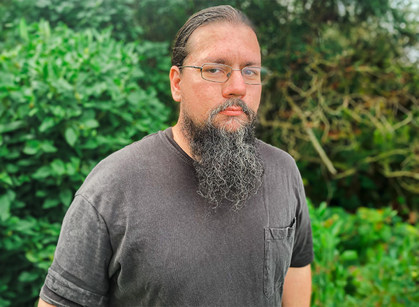 Michael George with glasses and  beard, shrubbery in the background.