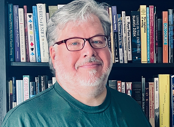 Mike Hill smiling at the camera with a shelf of books in the background.