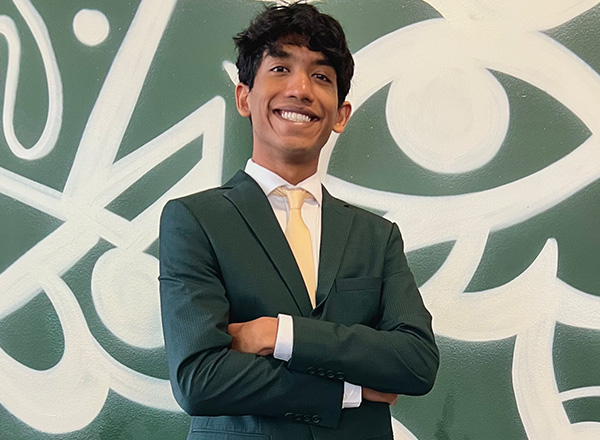 Sabit Islam wearing a green suit with yellow tie stands in front of a green and white background.