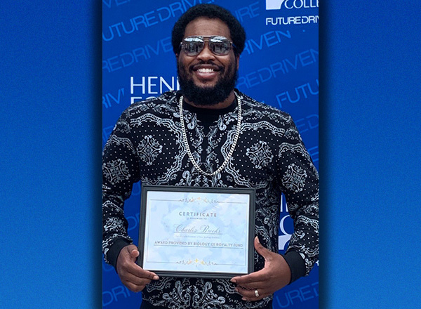 Charles Brooks smiling and wearing glasses, is holding a framed certificate in front of an HFC backdrop.