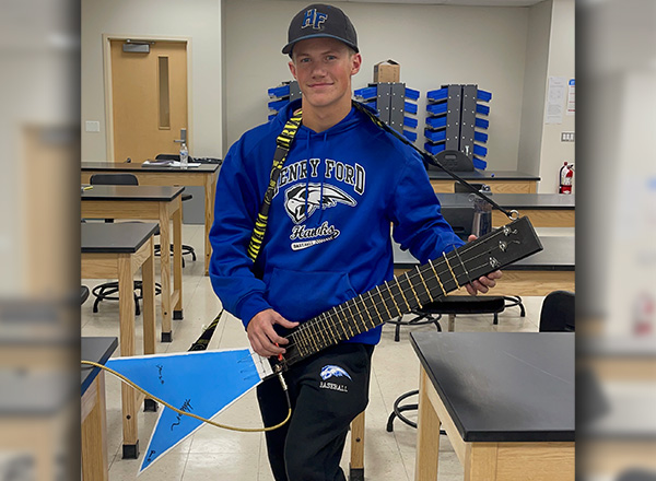 James Haranczak is wearing a blue Henry Ford Hawks hoodie and Henry Ford hat, while holding the electric guitar he made.