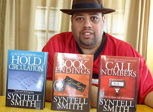 Les is wearing a black brimmed hat and red shirt, sitting behind three books