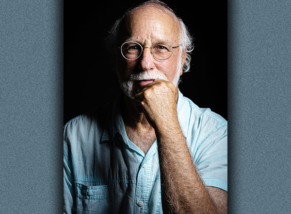 Headshot of Joen Geffen in thinker pose with black background. Joel has short white hair, wearing glasses, and a light blue shirt.