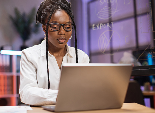 Woman in braids, glasses, and a white button down blouse working on a lap top.