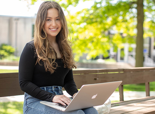 A young woman wearing a black shirt with long brown hair, working on her laptop outdoors