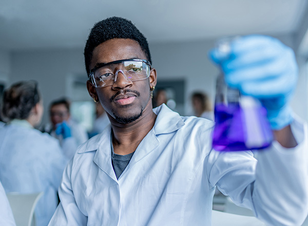 Young black man in a lab coat holding up a beaker of purple liquid.
