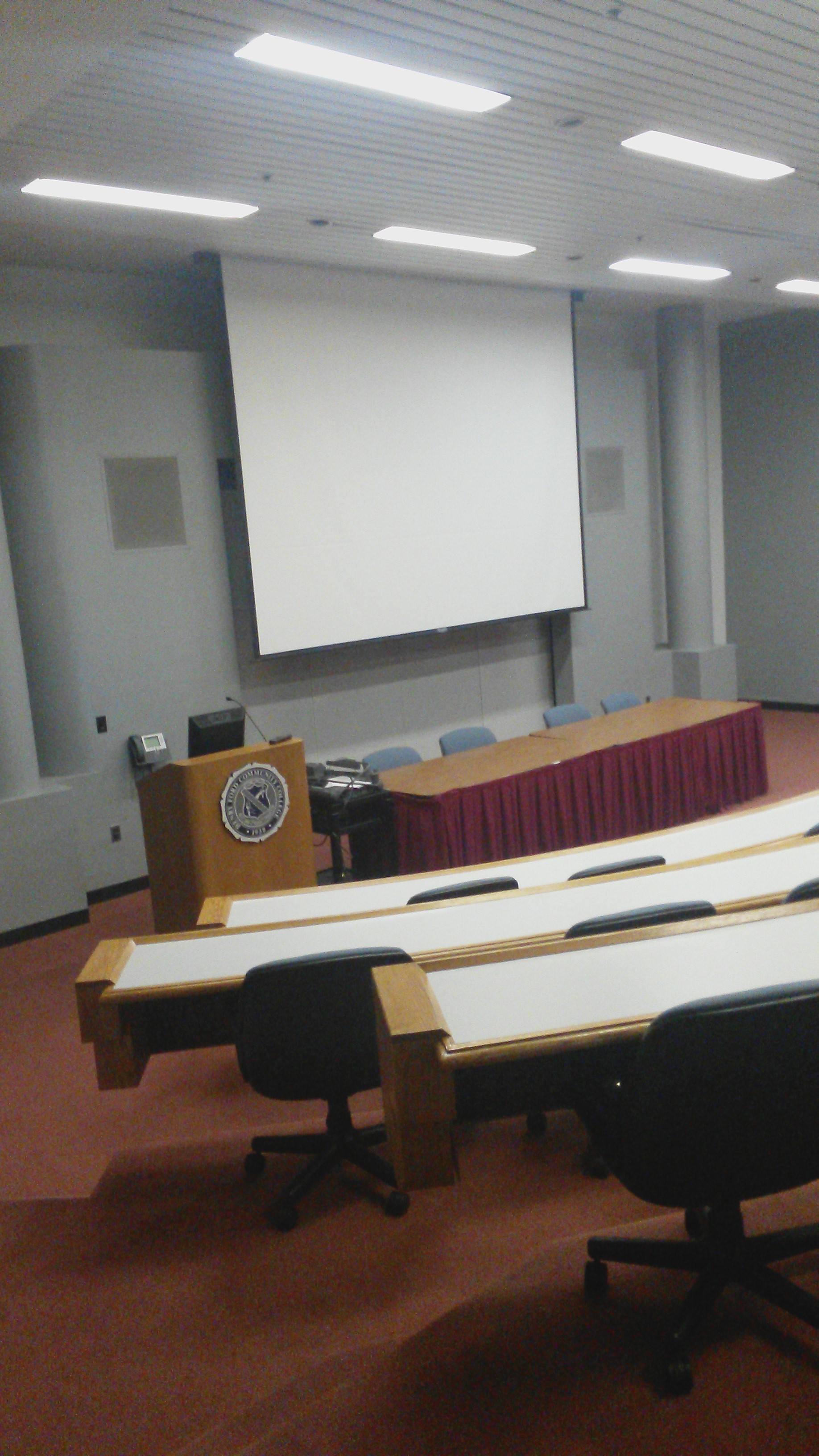 Photograph of the inside of Berry Auditorium, rows of auditorium seating and a large projector screen are visible, as well as a wooden podium next to a table with chairs in the front of the room in view of the "audience" 