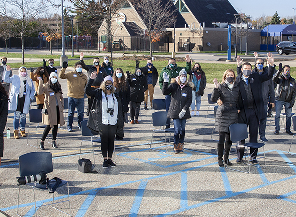 Members of the crowd wave to the camera, while staying 6 feet apart. 