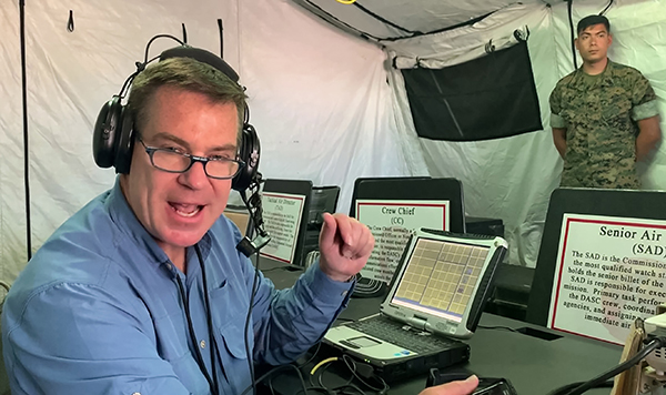 Radio host wearing headphones, with placards in front of him and a soldier in fatigues in the background.