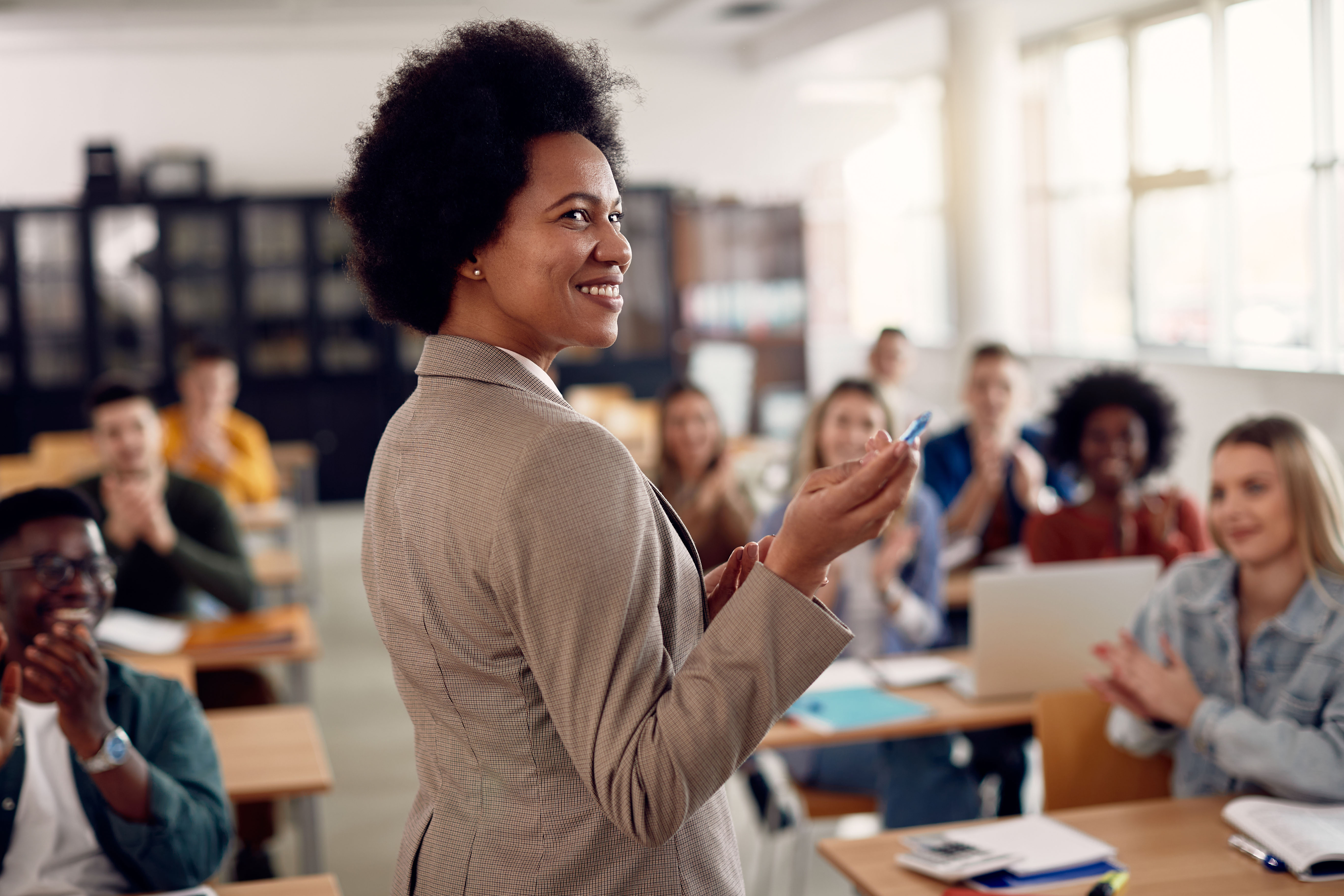 A teacher pointing in the front of class with students in the background.