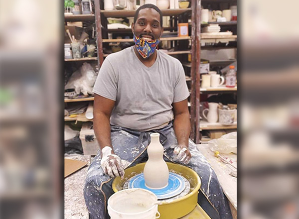 Centurium Frost wearing a mask and sitting in front of a pottery wheel with ceramics in the background.