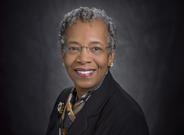 Saundra McGuire has short cropped grey hair, glasses, and wearing a black jacket with a gold and grey scarf. 