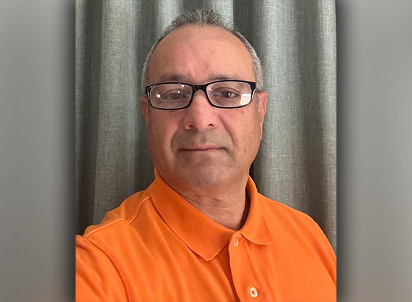 Sam Bazzi is wearing glasses and an orange polo with a grey curtain behind him. 