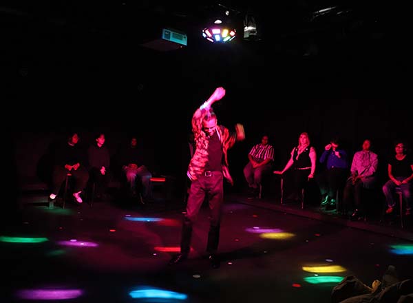 Theatre student dancing alone on a dark stage with colorful lights, for the production "Dear Harvey." The lone student is surrounded by other students sitting in chairs.