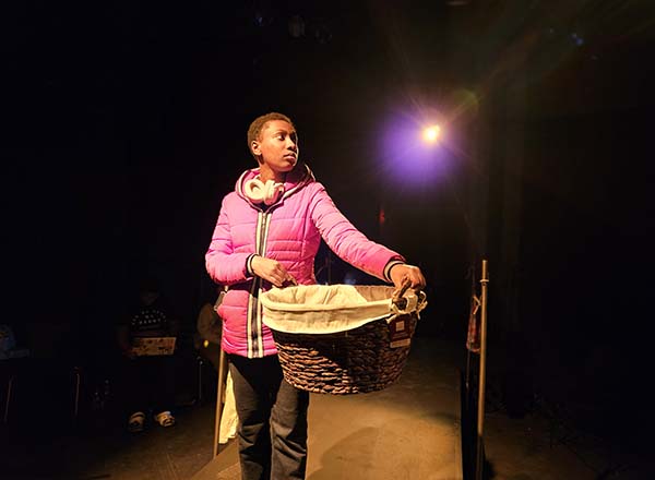 Theatre student in rehearsal, standing alone on dramatically lit stage, holding a large basket