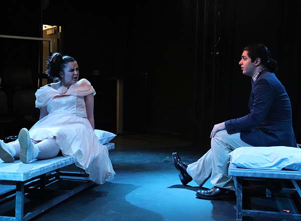 Two theatre students performing a scene from "Gruesome Playground Injuries," sitting across from each other on stiff cots. The student on the left looks upset; both are wearing semi-formal attire.