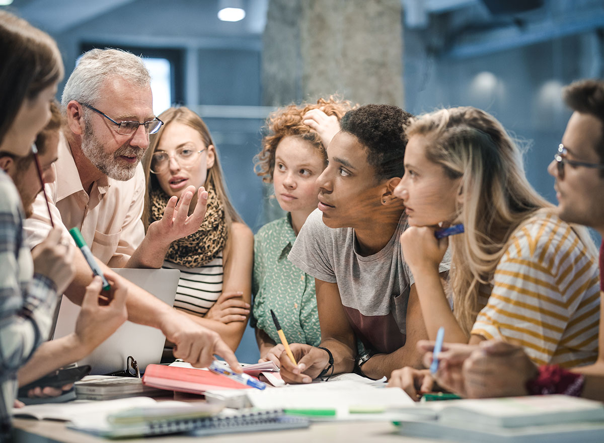 Teacher pointing to textbook while teaching a group of students, all standing around a small table
