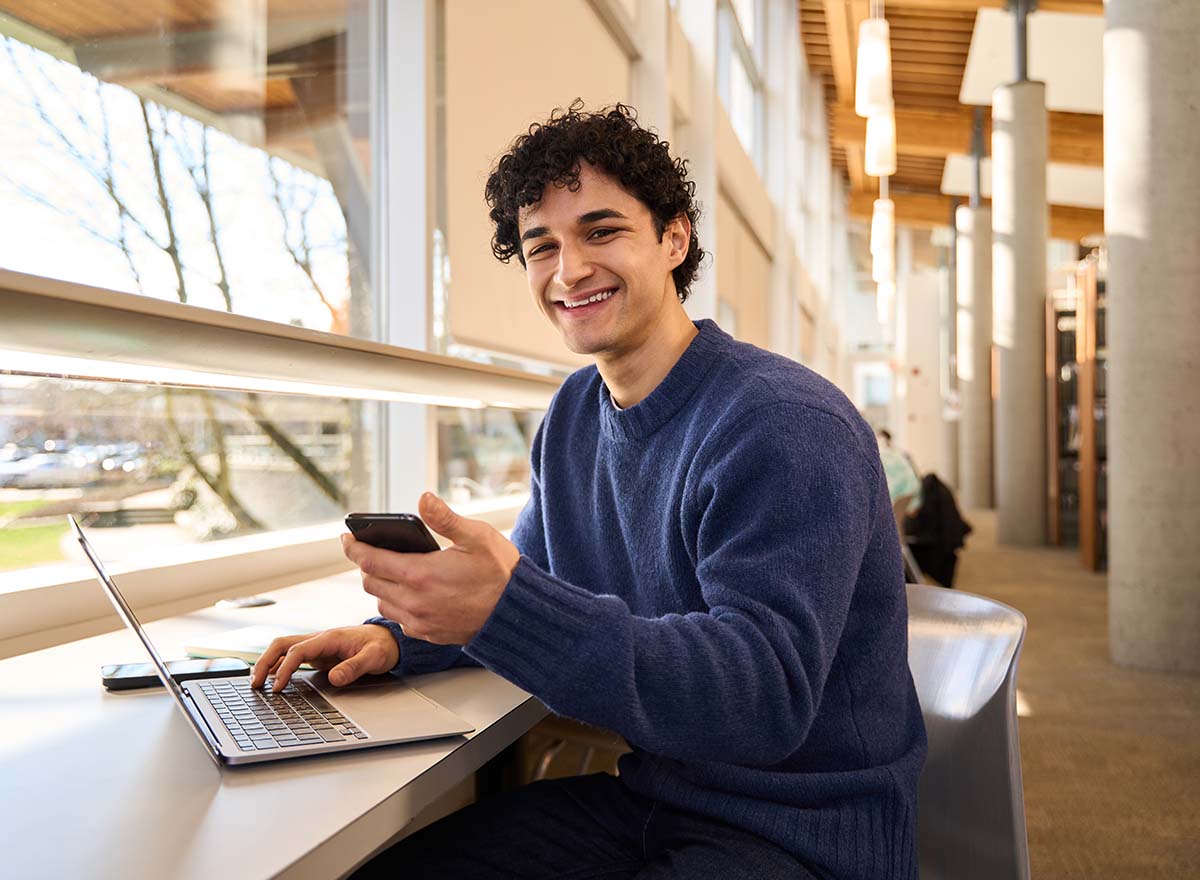 Smiling college student turned toward viewer, sitting at library desk with laptop