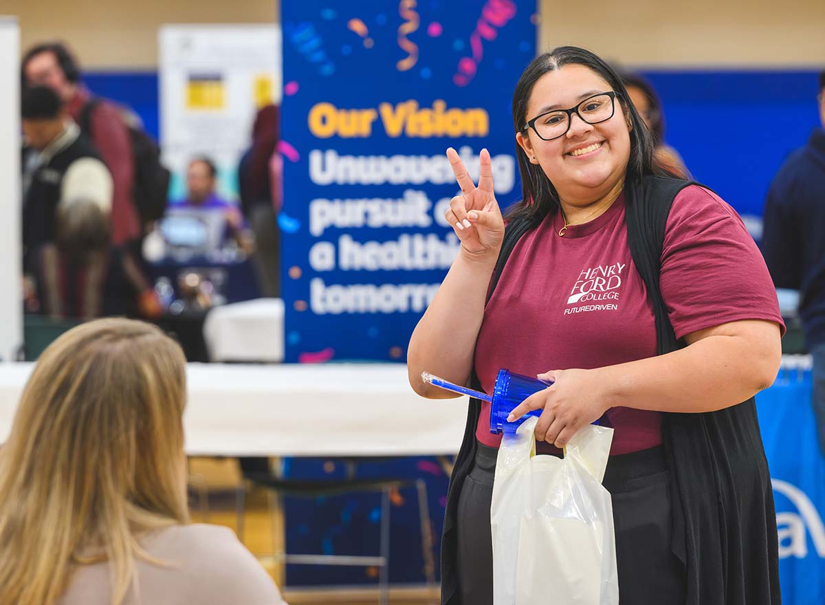 Smiling student standing at an HFC career expo booth, making a peace sign with their hand