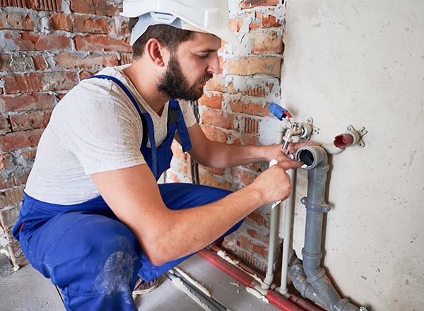 Plumber, wearing a white hardhat and blue overalls, applying sealant on apartment sewer pipe.