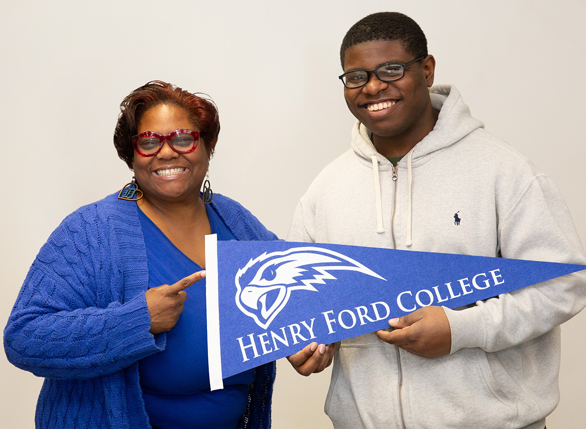 Staff member and student smiling and holding a Henry Ford College banner flag