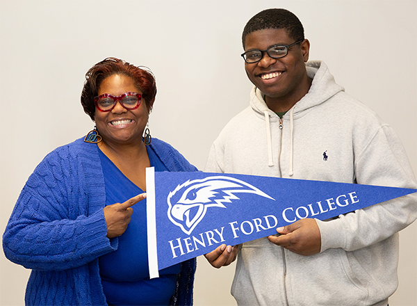 Staff member and student smiling and holding a Henry Ford College banner flag