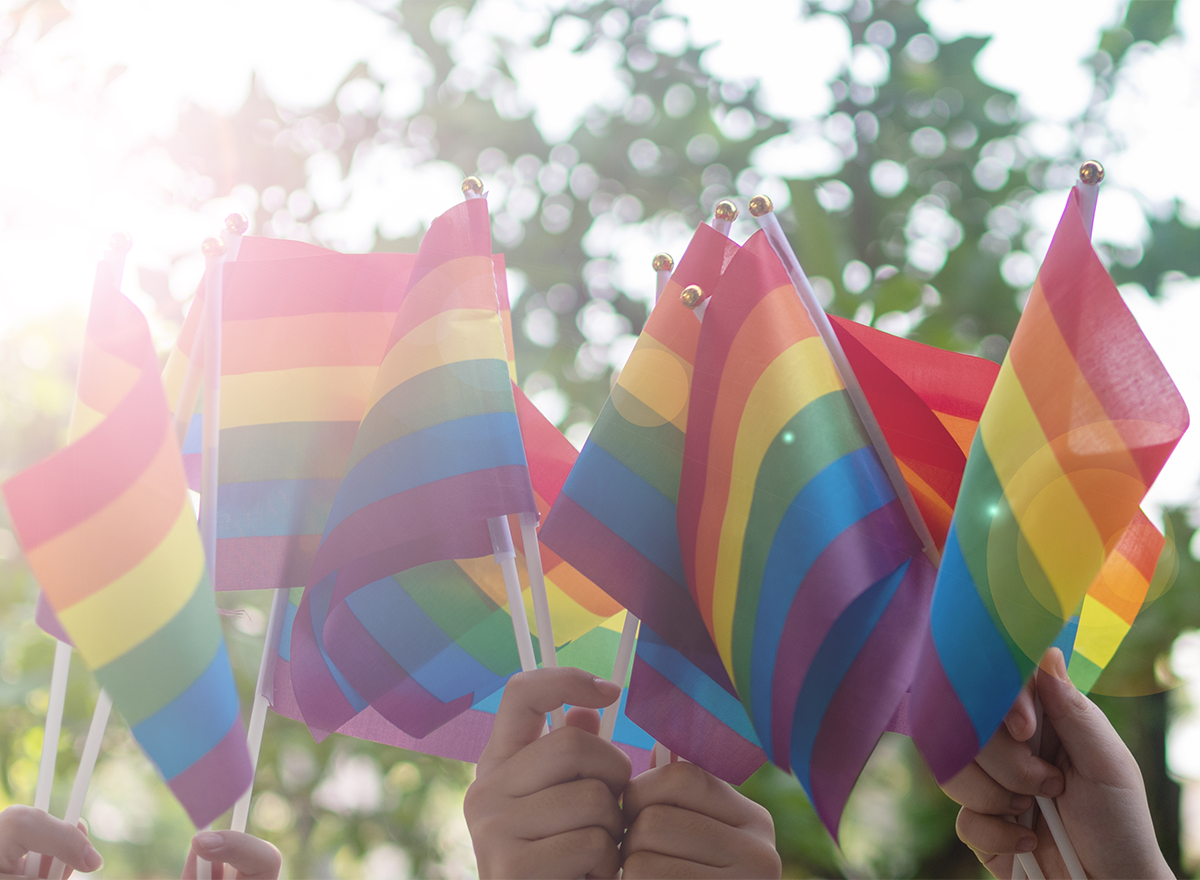 Five hands holding up small rainbow flags outdoors with trees in the background