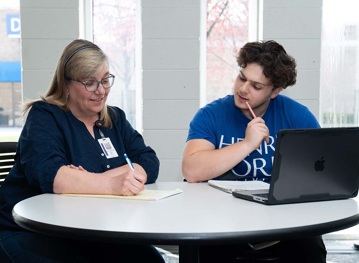 HFC student and instructor sitting at a table with a laptop, looking at a notebook.