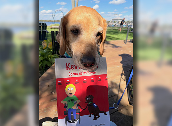 Trooper the service dog holds a copy of "Kevin Can! Cocoa Helps Kevin." Cocoa is based on Trooper.