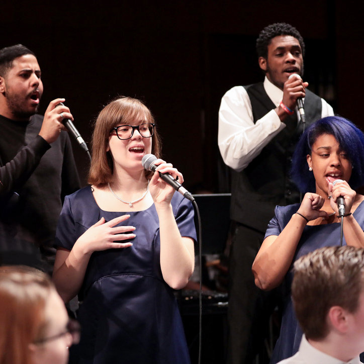 Four individuals singing as part of larger vocal ensemble on stage