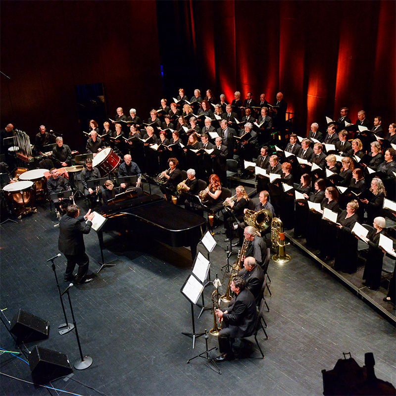 Aerial shot of full music ensemble performing under orchestral director, on stage with red velvet curtain background