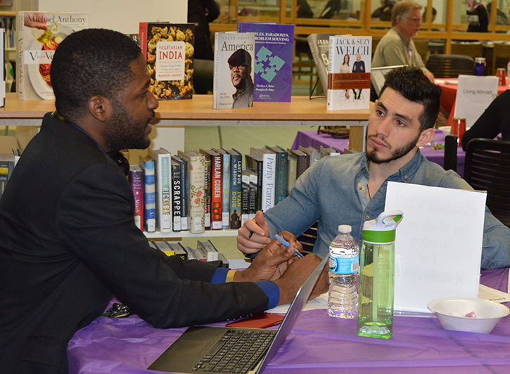 Two students sit at a table with shelves of books on display at library hosted "Human Library" event.