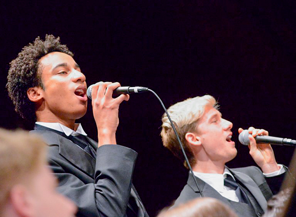 Two students singing into microphones, wearing black suits
