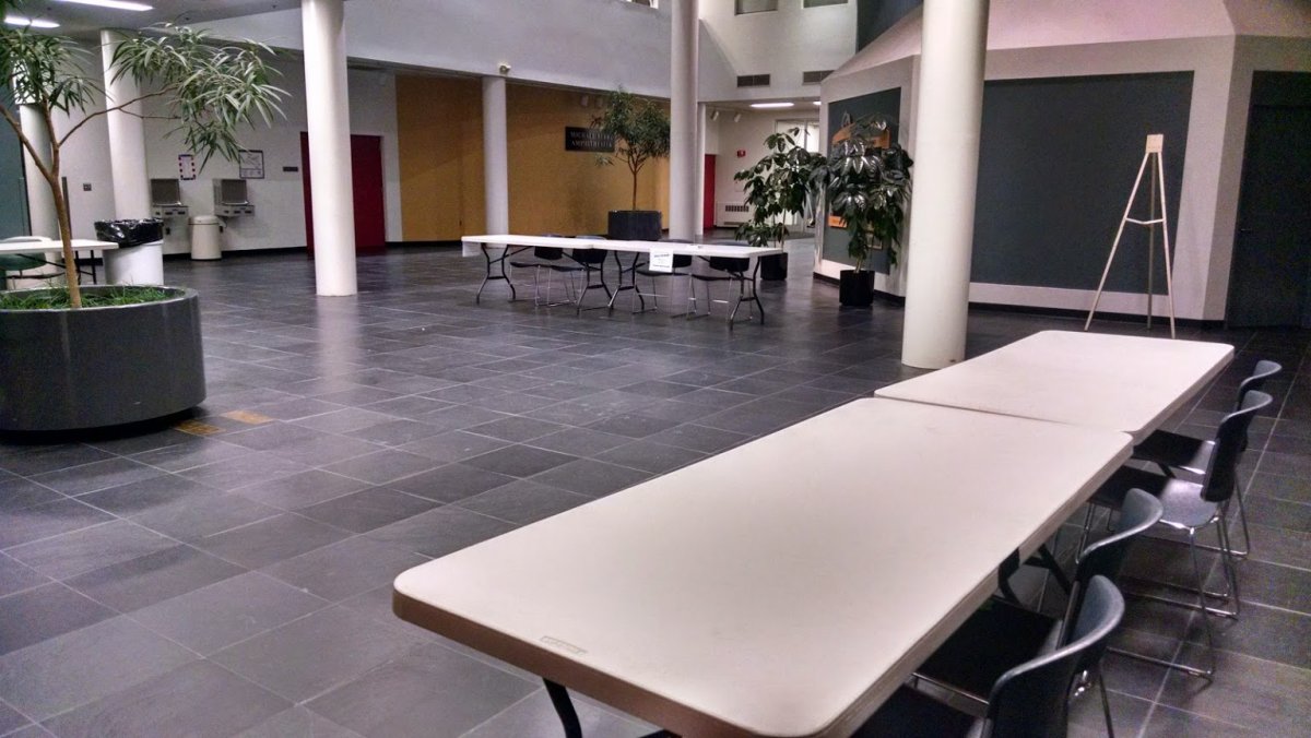 ASCC Atrium room L-111 pictured with tables and decorative plants; the room is large and has slate gray tiles, several white columns, and provides access to building exits and other rooms