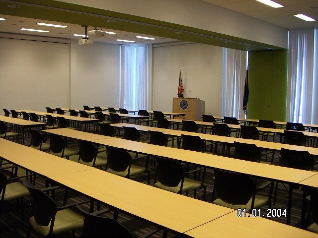 Inside of large room with several rows of long, rectangular tables and a podium at the front of the room where the chairs at the tables are facing