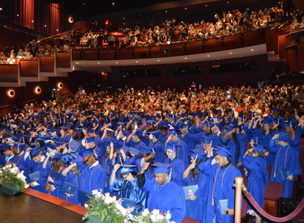 Hundreds of students wearing blue graduation gowns and caps stand in a theatre with their families near the back and on the balcony of the theatre applauding as the students move their tassels.