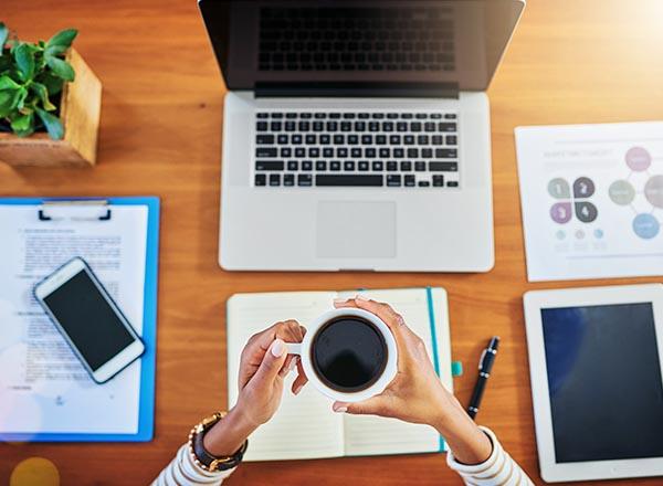 An image of a workspace with laptop, phone, tablet, journal, and coffee cup in hand.