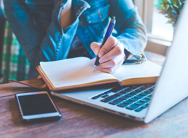 A person with pen in hand writing on a notebook and open laptop.