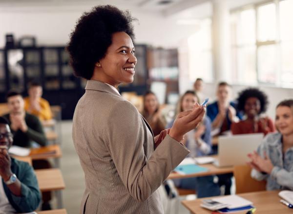 A teacher pointing in the front of class with students in the background.