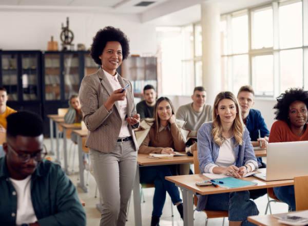 Image of a teacher standing, teaching a class, students seated in the class, interacting