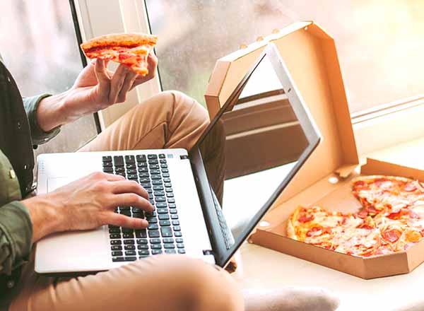 Student with a laptop computer, pizza slice in one hand, box of pizza in the background.