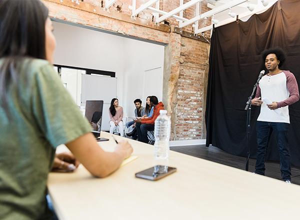 Photo of a student auditioning at a microphone for a drama teacher (other students are in the background).