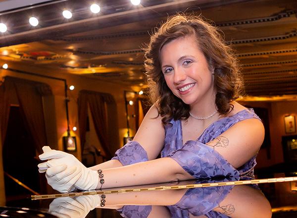 Olivia Van Goor leaning onto a piano top smiling.