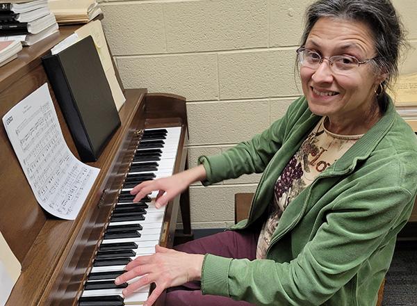 Gina is wearing a green sweater and glasses, playing the piano.