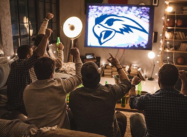 An image of people watching a large screen TV with the Henry Ford Hawks logo on it