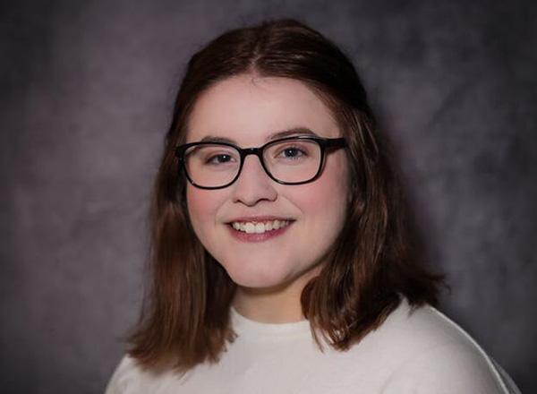 Cortney Markham wear black glasses and a white sweater, and has dark brown hair, smiling at the camera.
