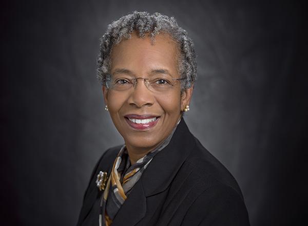 Saundra McGuire has short cropped grey hair, glasses, and wearing a black jacket with a gold and grey scarf. 