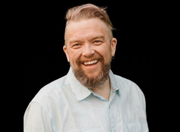 Dr. Adam Hazlett has blonde hair and facial hair, wearing a white button down with a black background.